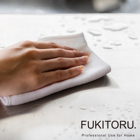 一隻手正使用白色超極細纖維擦拭布擦拭有水滴的銀色檯面，擦拭布質地柔軟，表面有網格紋理。