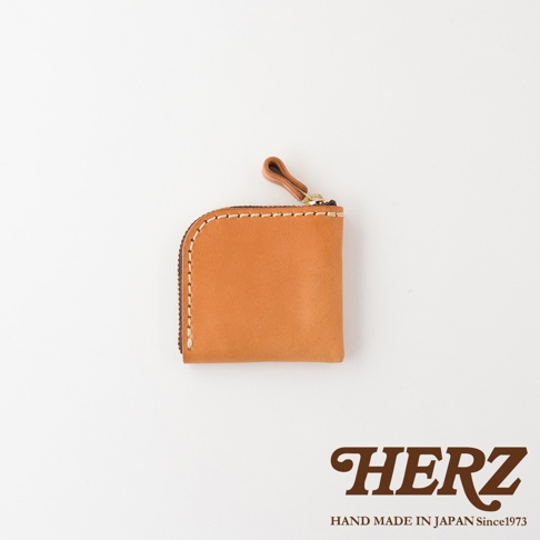 這是一款由日本品牌HERZ推出的職人手作皮革零錢包，外觀為淺棕色方形皮革，邊緣採用精細的粗線手工縫製，並設有頂部拉鍊與皮革拉環，下方印有HERZ品牌商標與產地說明，整體呈現簡約質感風格。