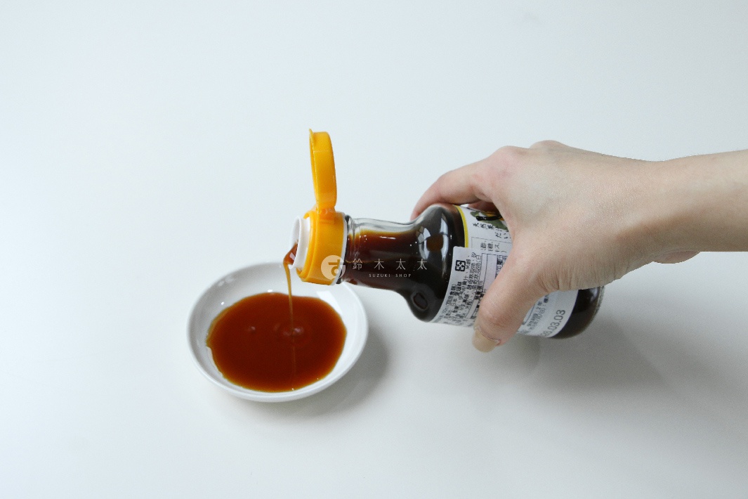 手持一瓶森文柑橘醋調味料，將琥珀色液體倒入白色小碟中。瓶身為深棕色玻璃，配有黃色翻蓋式瓶蓋。