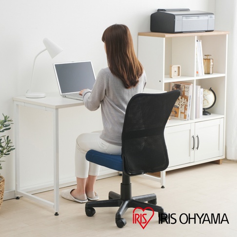 一位女子坐在藍色座墊的黑色網狀辦公椅上，她在一個白色書桌前使用筆記型電腦，桌上還有一盞白色檯燈。身後是一個白色開放式書架，上面放著文件、裝飾品和一個地球儀。