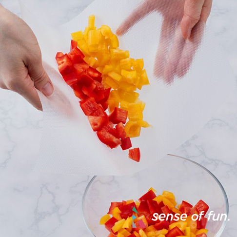 一雙手正拿著白色網狀紋理的捲筒式砧板保潔墊片，上面鋪有切塊的紅黃甜椒，準備倒入玻璃碗中。