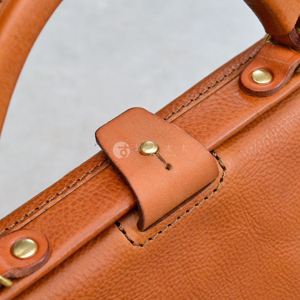 職人手作皮革Organ方形口金托特包，特寫展示包款的棕褐色皮革材質、精緻縫線與金屬卯釘細節，包含一個皮革束帶固定設計。