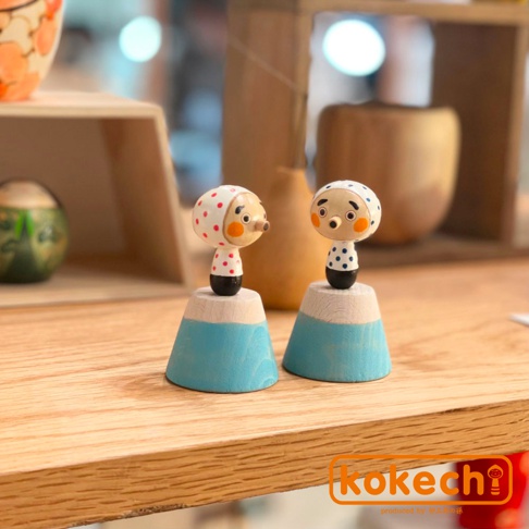 兩個木芥子開運擺飾，一個戴著紅色圓點頭巾，另一個戴著藍色圓點頭巾，均立於淺木色圓柱上，圓柱下為藍色錐形底座，表情生動可愛。