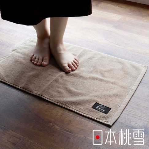 一塊淺棕色、觸感柔軟的毛巾材質浴墊，上面有黑色的品牌標籤，一雙赤腳踩在其上，置於木紋地板上。