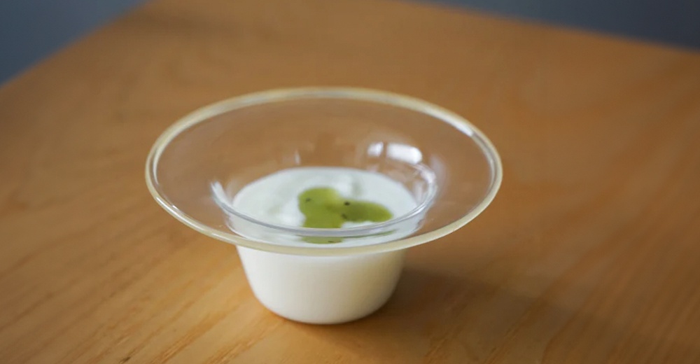 一個白色圓形點心碗，上面盛有白色優格和綠色奇異果醬，外層套著一個透明玻璃盤。