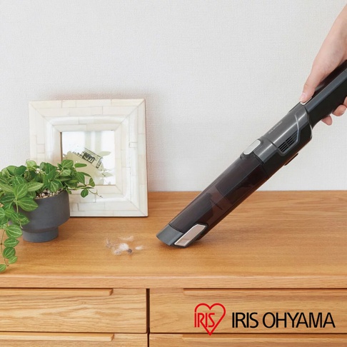 手持式深灰色充電吸塵器，配有透明集塵筒，正在清理桌面上的灰塵碎屑。產品上方印有「IRIS OHYAMA」品牌標誌。