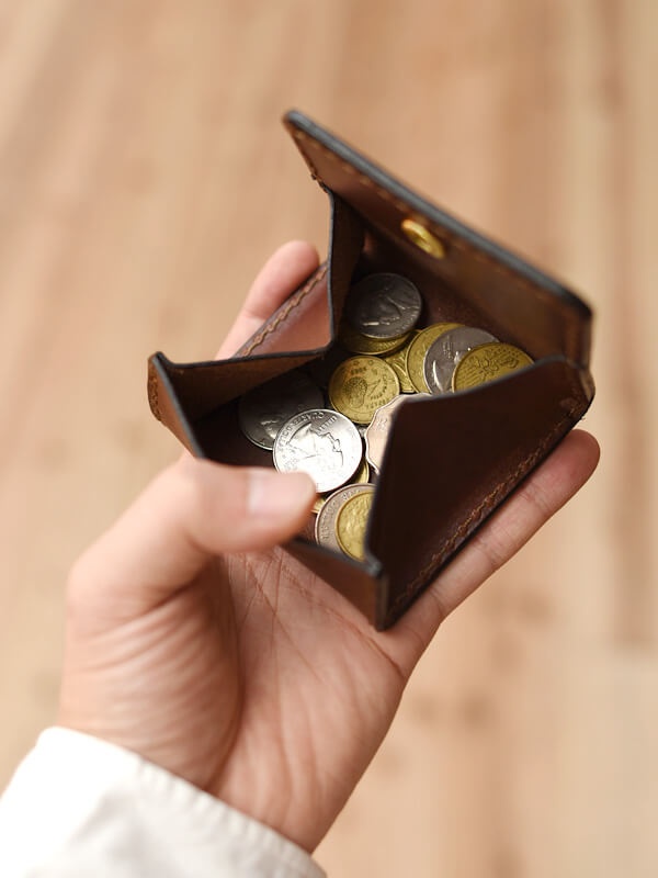 一只手拿著一個深棕色皮革製的箱型收納零錢包，零錢包內裝有數枚銀色和黃銅色的硬幣。