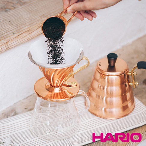銅製V60濾杯，帶有金色把手，置於玻璃咖啡壺上，旁邊有一個銅製手沖壺。
