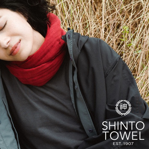 一位女性臉部特寫，身穿紅色針織圍巾、灰色上衣和深灰色外套，背景為乾枯的草叢。