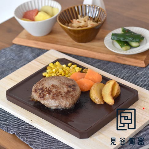 HAI減煙火山灰烤盤，長方形，深棕色，材質為火山灰陶。盤上盛有漢堡排、玉米粒、胡蘿蔔塊和馬鈴薯塊，整體呈現日式餐點風格。