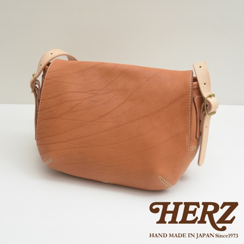 棕色皮革製成的郵差包，帶有可調節的淺色皮革肩帶和金屬扣環。包身有自然的皮革紋理和手工縫線細節。