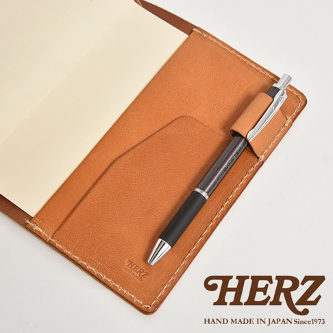 皮革手帳書套內夾著一支筆，左側為空白內頁，右側有筆插與口袋，品牌字樣HERZ印在皮革上。