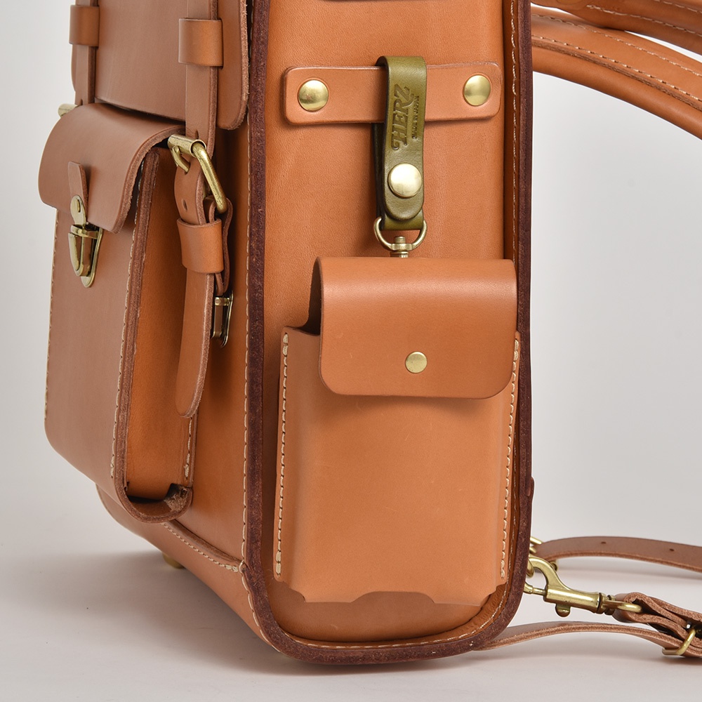 職人手作皮革經典學院風後背包，採用鞣製皮革材質，呈現自然棕色調，背包側面有雙層口袋設計，並附有軍綠色皮革吊牌及黃銅五金配件。