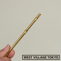 一根黃銅製成的細長線香架，表面呈現金屬光澤，並帶有仿竹節的紋路，一端為開放式管狀設計，由一隻手持握。