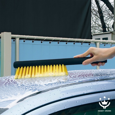 手持黑色軟柄洗車刷，黃色刷毛沾著泡沫，正清潔著覆蓋著泡沫和水珠的汽車車頂。