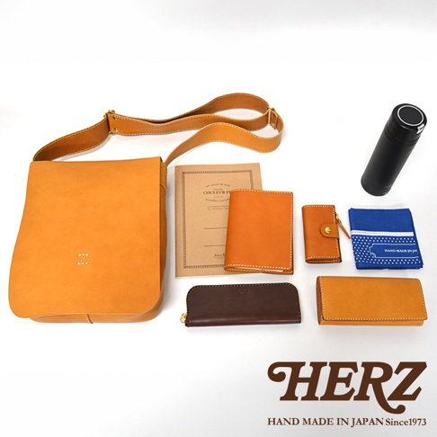左側為駝色皮革斜背包，上方擺放著一本筆記本、兩個皮革皮夾，下方有咖啡色長夾及一個駝色皮夾，最右邊為黑色保溫瓶和藍色手帕，下方壓印有HERZ品牌標誌及HAND MADE IN JAPAN Since1973字樣。