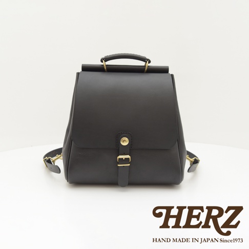 黑色皮革後背包，具有屋瓦造型翻蓋，搭配金色釦環和手提把，風格簡約。