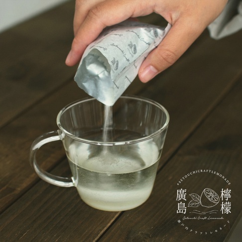 一隻手正將廣島檸檬茶粉末倒入透明玻璃杯中的清水裡，杯中裝有清澈的飲用水。畫面右下角帶有廣島檸檬的圖案及 Setouchi Craft Lemonade 的品牌標誌，整體呈現出清新、簡約的沖泡過程。
