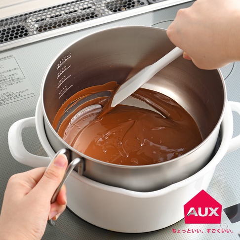 附手把的刻度攪拌鍋，內鍋為不鏽鋼材質，盛裝著融化的巧克力。白色外鍋，鍋壁有刻度標示。一支白色刮刀正在鍋中攪拌巧克力。