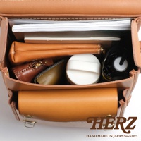 職人手作皮革小學生書包內部，可見文件、筆袋、化妝包、鑰匙包、綠色卡夾、白色圓形小物及黑色雨傘，皮革為自然棕色。