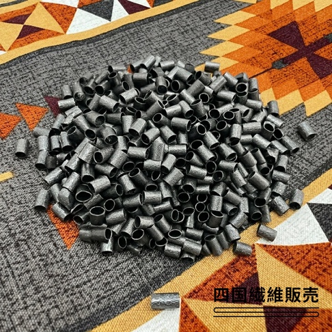一張擺放在民族風格織物上的備長炭軟管填充物，這些灰黑色的小圓柱體呈現粗糙質感，散落在布面中心，畫面右下角帶有四國纖維販賣的文字。