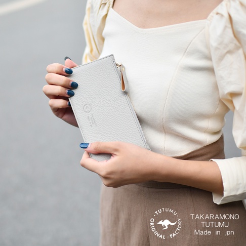 一位女士手持淺灰色皮革拉鍊長夾，夾子上印有品牌標誌和文字 TAKARAMONO TUTUMU Made in jpn