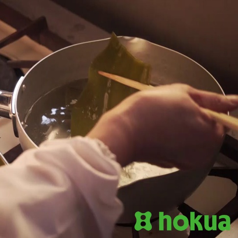 職人槌目雪平鍋，鍋身為銀色鋁製，鍋內有水正在加熱，一塊綠色海帶由木筷夾住，鍋具旁有白色的廚師服。