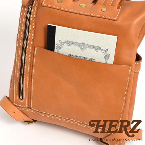 棕色皮革後背包，設有雙口袋，其中一個口袋內插有一本寫著「NOTE BOOK」的筆記本。包款使用粗獷的縫線，並有金屬拉鍊和铆釘裝飾。