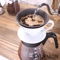 白色陶瓷濾杯架上，黑色濾杯中正注入熱水，咖啡粉與泡沫混合，下方為帶有刻度的透明玻璃咖啡壺，整體置於木紋桌面上。