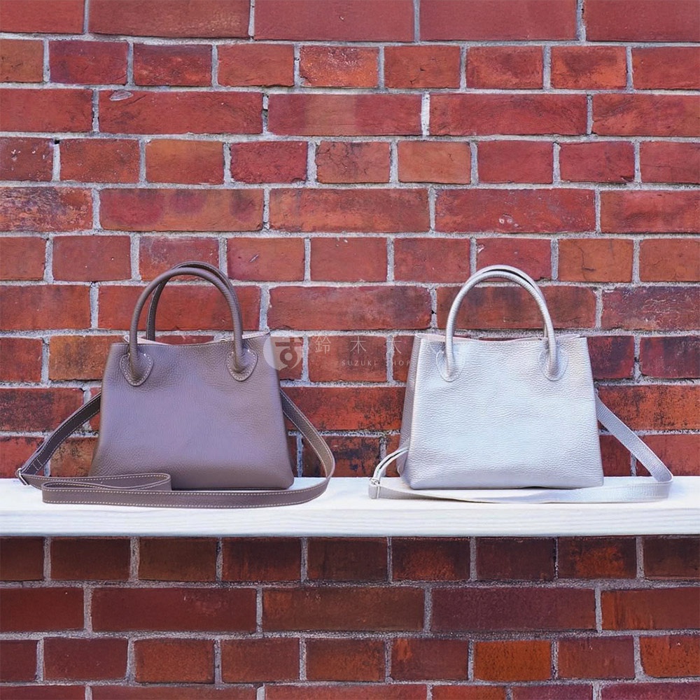 兩款京都職人手作兩用手提托特包，一款為淺灰色，另一款為銀白色，皆為皮革材質，配有短提把與長背帶，置於紅磚牆背景下的白色層板上。