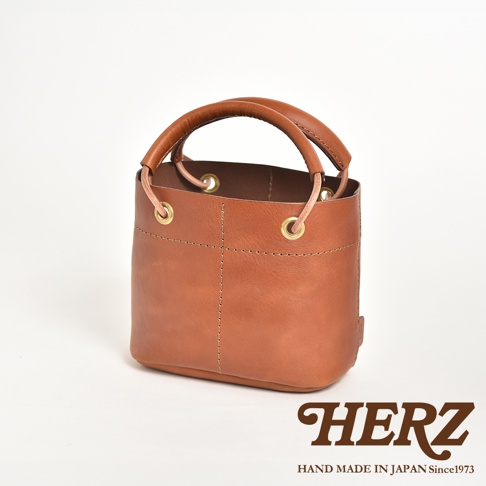 棕色皮革製成的圓筒形午餐袋，配有加厚提把，袋口有銅環及束繩設計，整體採用職人手作縫線工藝。