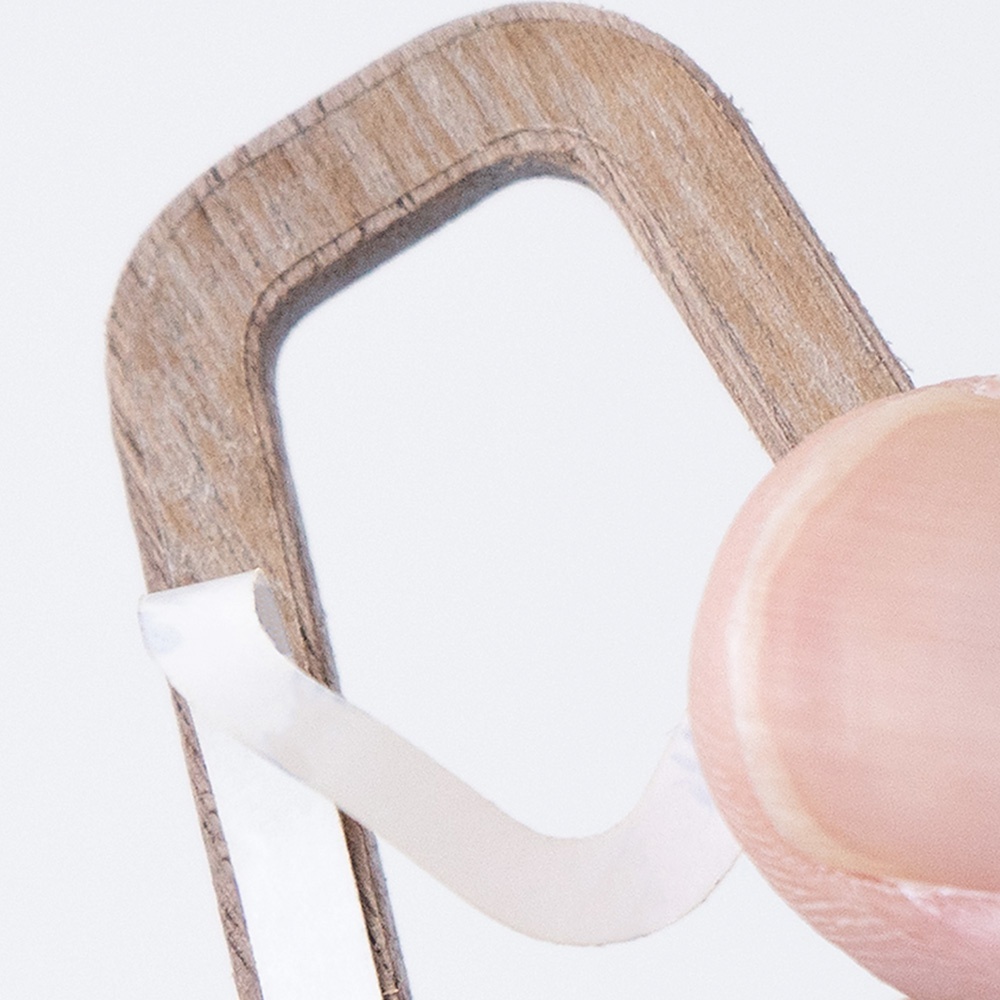 特寫鏡頭顯示一個淺棕色木製標示牌的局部，邊緣有木紋細節，一塊白色背膠正被從標示牌背面撕下。