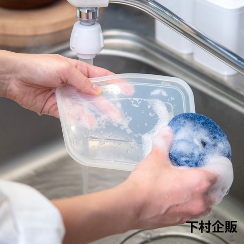 在廚房水槽邊，一個人正使用藍色圓形清潔海綿清洗透明的塑膠食品保存盒，水龍頭流出的水正沖洗著盒子，泡沫覆蓋在海綿與盒蓋表面，右下角標示著下村企販品牌字樣。