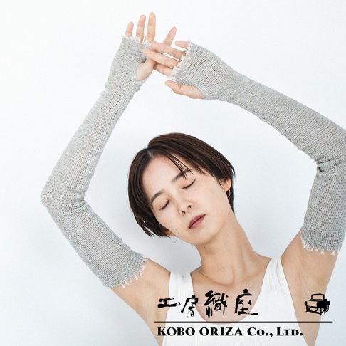 模特佩戴一對淺灰色、長及手臂的針織遮陽袖套，袖套採用網狀織法，指尖處露出，袖口有流蘇裝飾，材質輕柔。