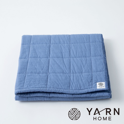 一條藍色、以格紋縫製的浴巾，由脫脂棉混紗製成，質地柔軟，邊緣有品牌標誌。