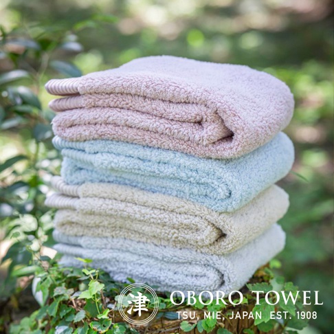 堆疊的毛巾，由左至右顏色為淺粉色、淺藍色、米白色和淺灰色。毛巾質地蓬鬆，邊緣有條紋裝飾。背景為綠葉模糊的自然環境。