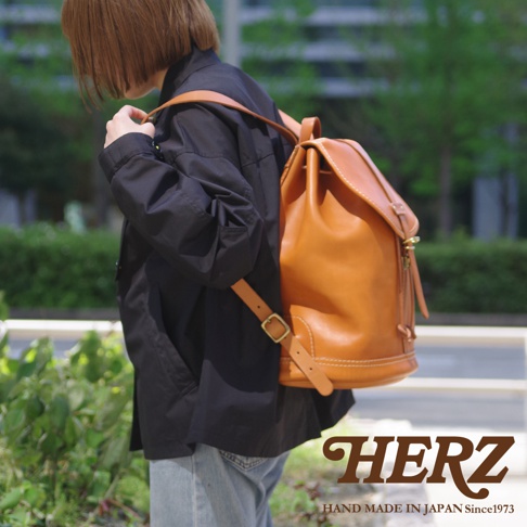 身穿黑色外套的女性背著一個棕褐色皮革單扣束口後背包，包款有明顯的縫線細節與金屬扣環。