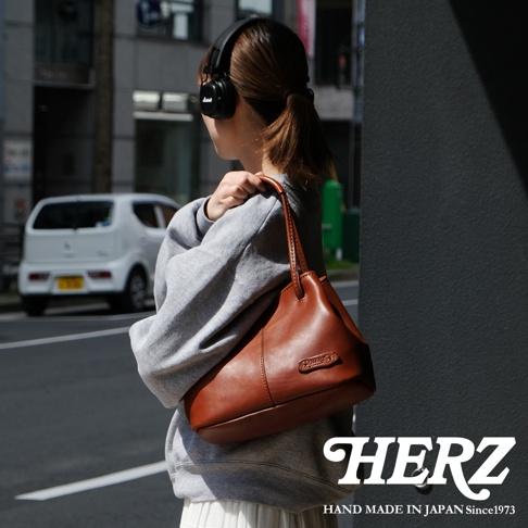 一位戴著 Marshall 耳機的女士，身穿灰色連帽衫，斜挎一個棕色皮革托特包，包上有 HERZ 品牌標誌，她站在建築物陰影下的街道旁。