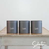 三個灰色圓筒形包裝盒，分別印有白色「Sodateru TOWEL」字樣，整齊排列在舊木桌上，背景為白色。