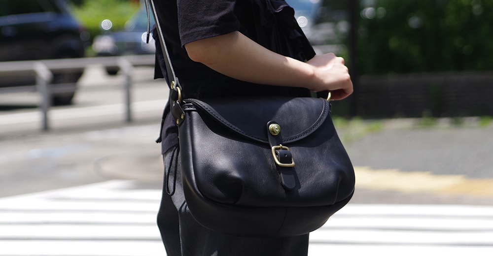 一個身穿黑色上衣的人，斜背著一個黑色皮革馬鞍包，包上有一個黃銅色金屬扣。