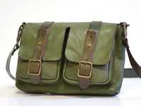 綠色皮革雙口袋肩背包，帶有兩個翻蓋，每個翻蓋都配有皮帶和金屬扣。包包有深色皮革飾邊和肩帶，並採用手工縫線。