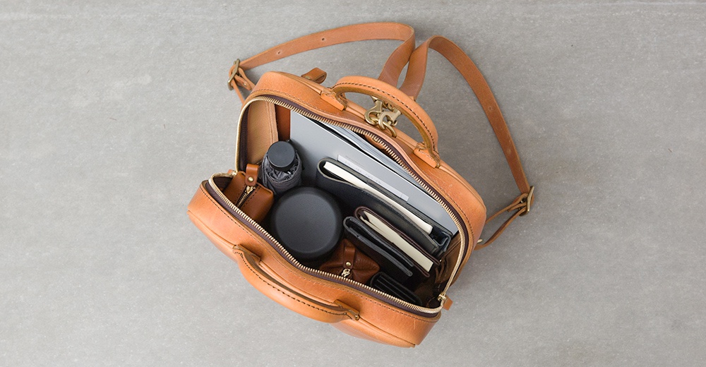 俯視視角，一個打開的棕褐色皮革後背包，裡面裝有筆記本、文件夾、傘、錢包和化妝包。