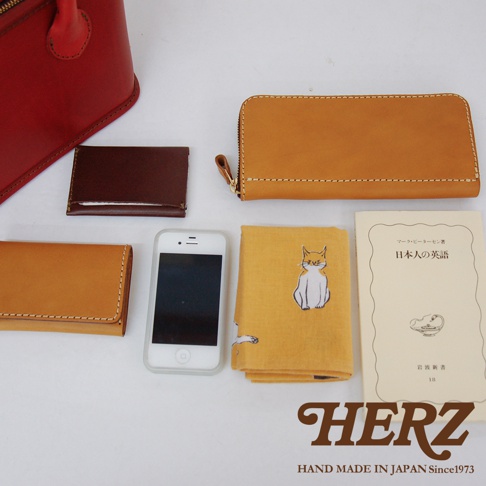 職人手作皮革輕巧散步手提包 Q-1 (共5色)：包含一個紅色皮革手提包，一個深棕色皮革名片夾，一個焦糖色皮革長夾，一個焦糖色皮革短夾，一個白色智慧型手機，一本印有橘貓圖案的黃色布巾，以及一本日文書籍。