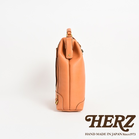 側面視角的日本HERZ職人手作皮革圓釦後背包，呈現經典的淺棕色植鞣革質感，袋身造型立體扎實，細節處可見黃銅五金零件與精緻的縫線工藝，底部右下方印有品牌名稱與創立年份標誌。