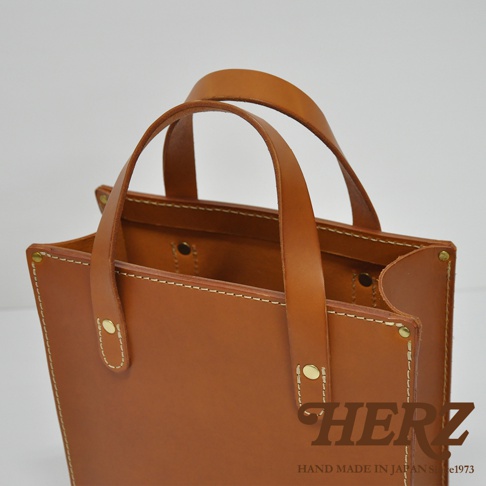 職人手作皮革直式托特包，採用天然皮革材質，呈現溫潤的焦糖棕色。包款設計簡潔，有兩個皮革提把，以白色縫線和金色銅釦裝飾，整體風格樸實耐用。