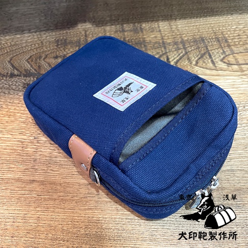 深藍色石洗帆布斜背手機包，正面有方形白色貼布標籤，標籤上有日文圖案與文字，包包側面有棕色皮革裝飾，拉鍊開口，材質厚實耐用。