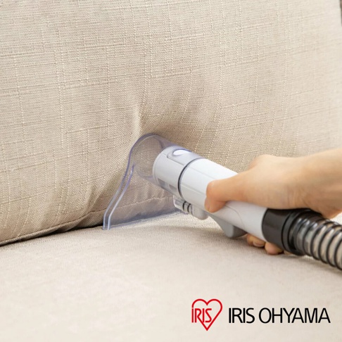 手持白色自動給水織物清潔機的清潔頭，正對著米色布質沙發的縫隙進行清潔，清潔頭為透明塑膠材質，機身為白色與黑色相間。