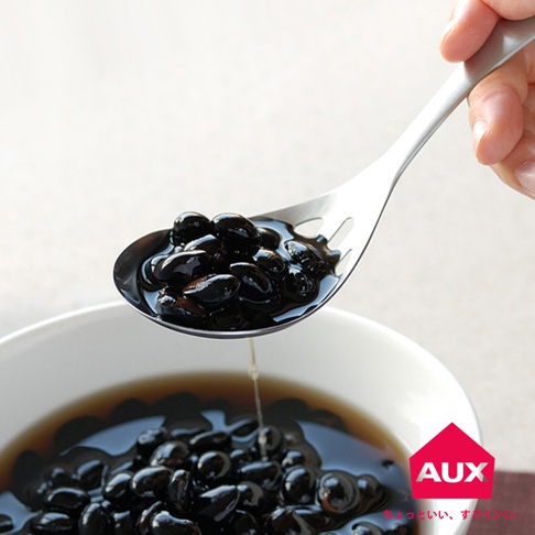 銀色金屬濾水菜匙，盛著大量光滑的黑色豆子，湯汁從匙孔中瀝出，下方為裝有黑色豆子的白色碗。