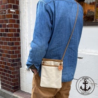 米白色帆布散步包，搭配棕色皮革飾邊與細肩帶，側面有一個圓形皮革吊牌，上面印有船錨圖案。