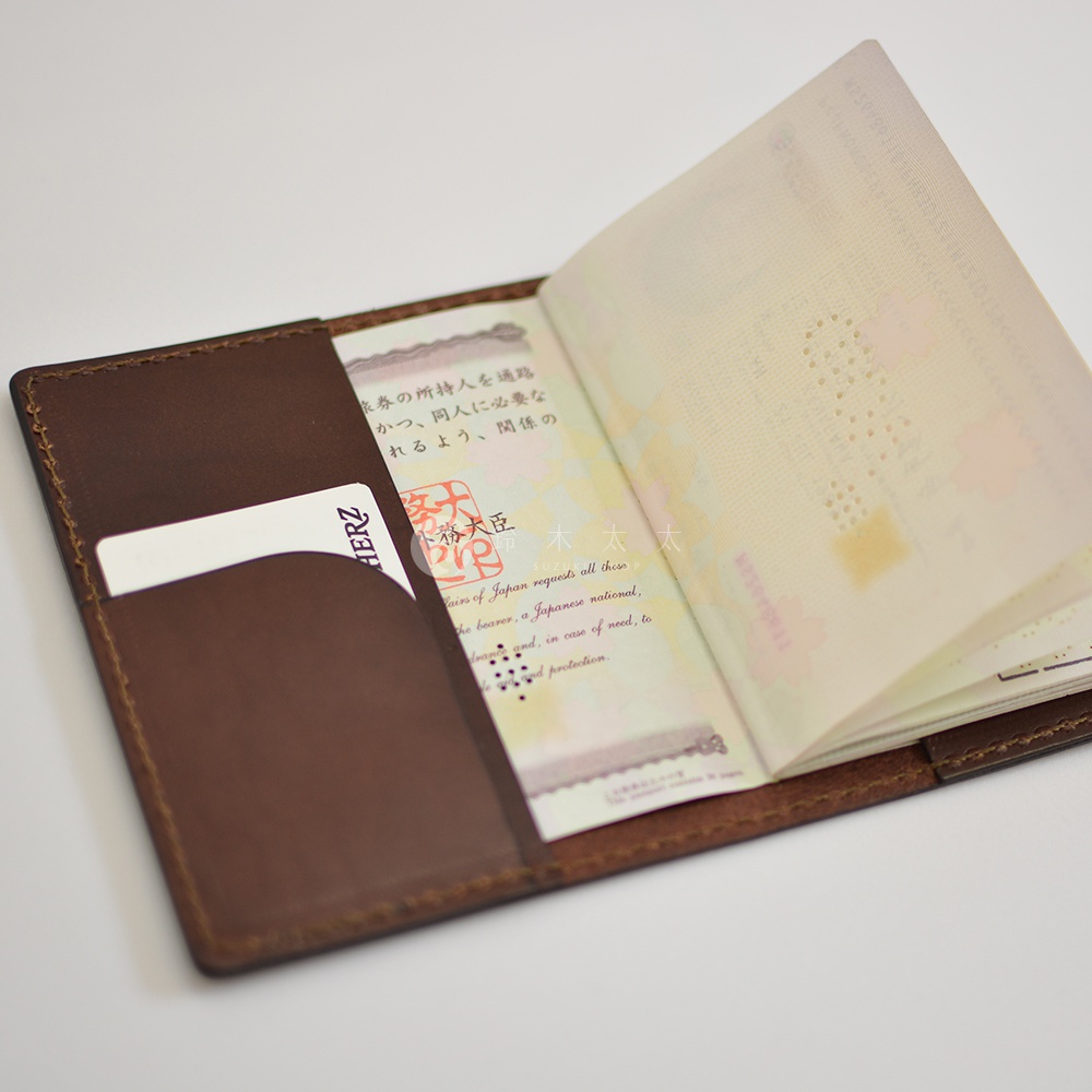深棕色皮革製成的職人手作經典護照套，內含卡槽，左側可見部分白色卡片。護照套打開，露出其中一頁的日文及英文文字，以及一頁印有日文圖案的護照內頁。